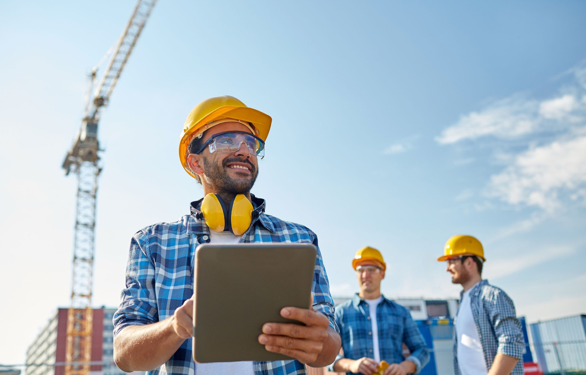 Bauarbeiter mit Schutzhelm, Gehörschutz und Schutzbrille hält ein Tablet auf einer Baustelle. Im Hintergrund weitere Arbeiter mit Helmen, ein Kran und Container